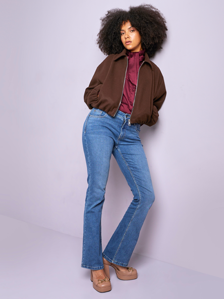 vrouw leunend tegen muur met flared fit jeans en een bruin jasje